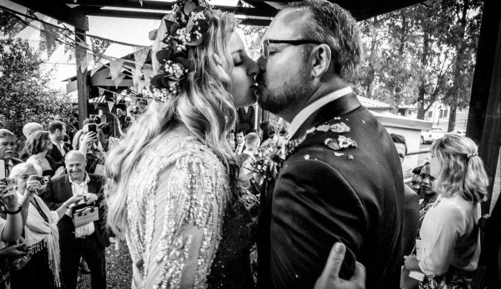 A black and white photo of a bride and groom kissing while the wedding guests watch in the background.