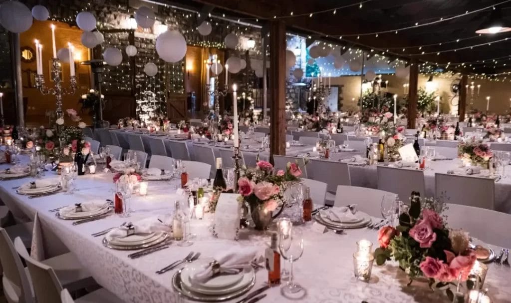A wedding reception set up with white table cloths, flowers and fairly lights next to grey stone walls.