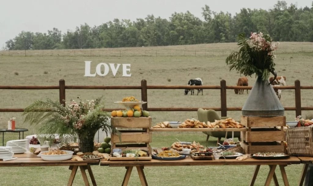 Outdoor buffet table set with various dishes and flowers, with a field, grazing cows, and a large "LOVE" sign in the background.
